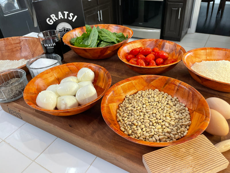 A wooden countertop displays bowls filled with ingredients, including mozzarella balls, cherry tomatoes, spinach, white beans, shredded cheese, flour, seasonings, eggs, and a glass of water, ready for cooking.