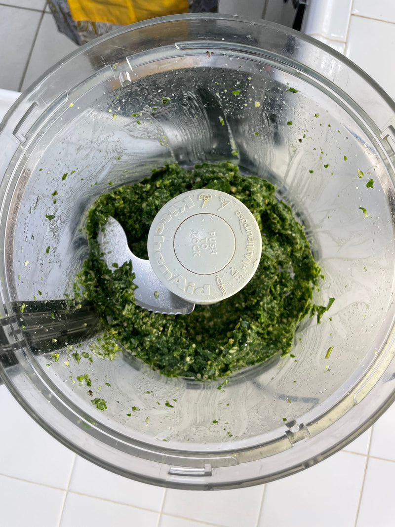 A food processor containing freshly blended green pesto sits on a white kitchen counter. The pesto is visibly textured with chopped herbs and ingredients around the blade.