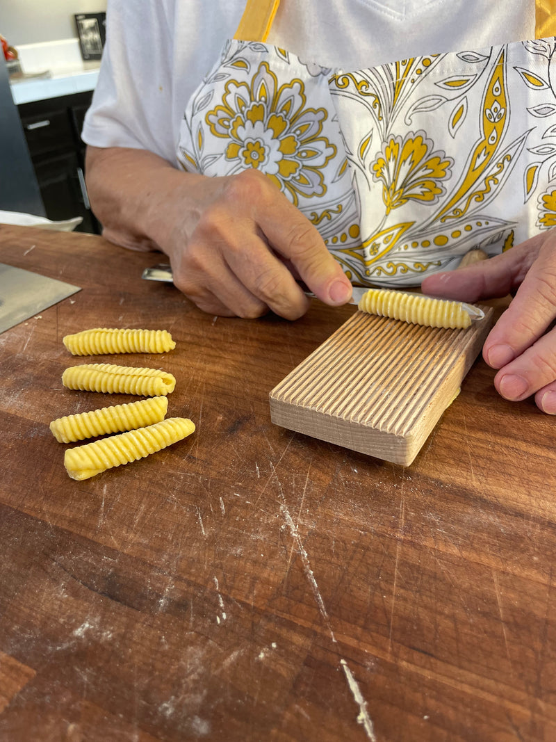 Close-up of a person shaping ridged pasta with a wooden board on a wooden surface. Several pieces of pasta are finished, and the person is wearing a white apron with a yellow floral pattern.