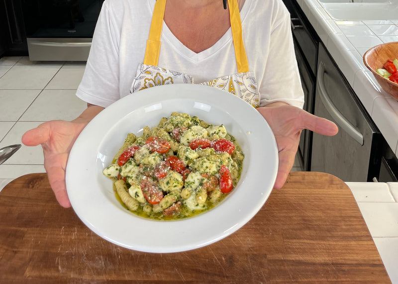 A person in a white shirt and yellow apron holds a white bowl of gnocchi with pesto sauce, cherry tomatoes, and grated cheese over a wooden countertop in a kitchen.