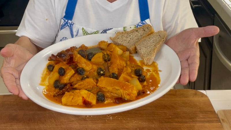 A person in a white shirt and blue apron holds a plate with chicken stew, potatoes, black olives, and tomato sauce, garnished with two slices of whole grain bread.