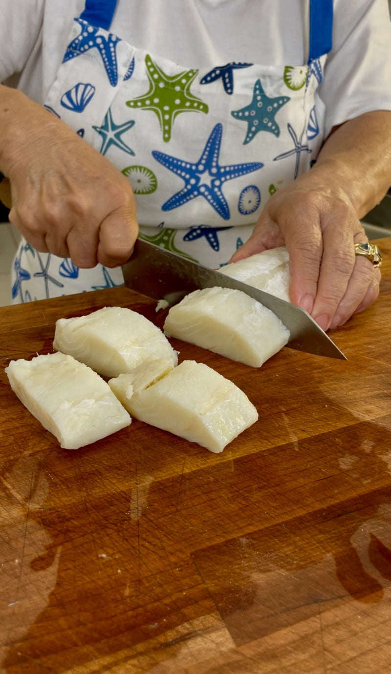 A person wearing a starfish-patterned apron slices white fish fillets into chunks on a wooden cutting board with a large kitchen knife.