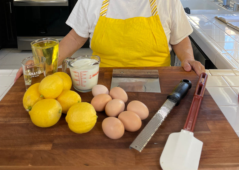 A person wearing a yellow apron stands behind a wooden counter with lemons, eggs, a cup of milk, a glass of sugar, oil, a zester, a spatula, and a dough scraper arranged in front of them.