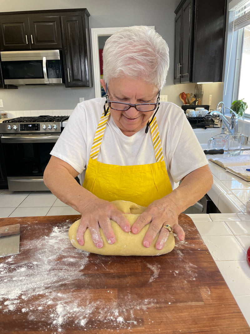 An older woman with short white hair and glasses kneads dough on a floured wooden surface in a kitchen. She wears a yellow apron over a white shirt and is smiling as she works.