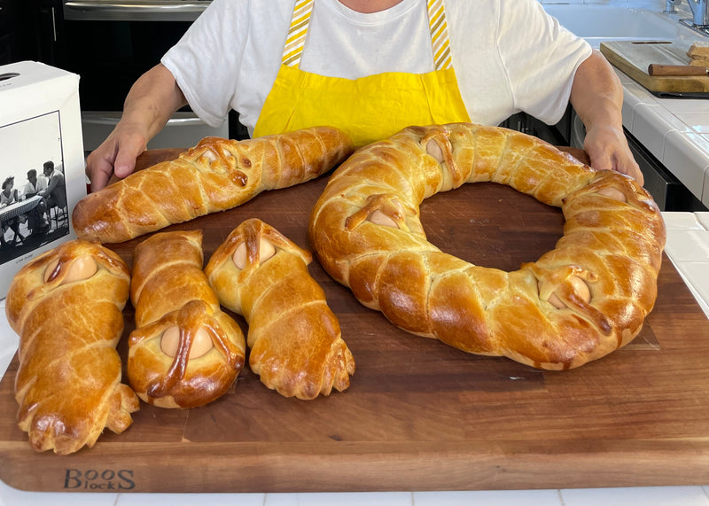 A person in a yellow apron holds a wooden board with three bread loaves shaped like swaddled babies and a large, braided ring-shaped bread in a bright kitchen.