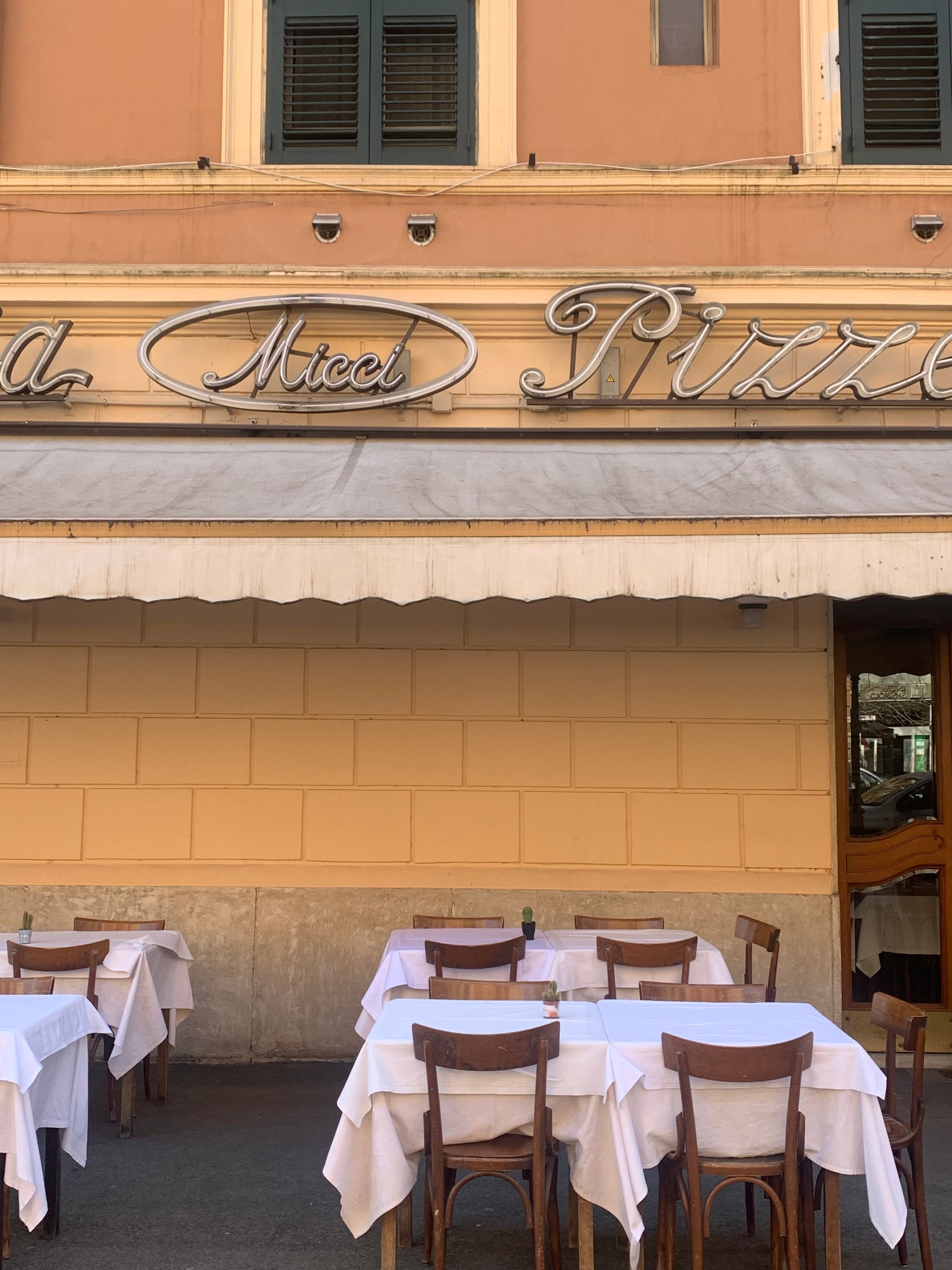 Outdoor seating area of an Italian restaurant with wooden chairs and tables covered in white tablecloths, beneath a cream awning. The building has a yellow facade and a sign with cursive writing above the entrance.