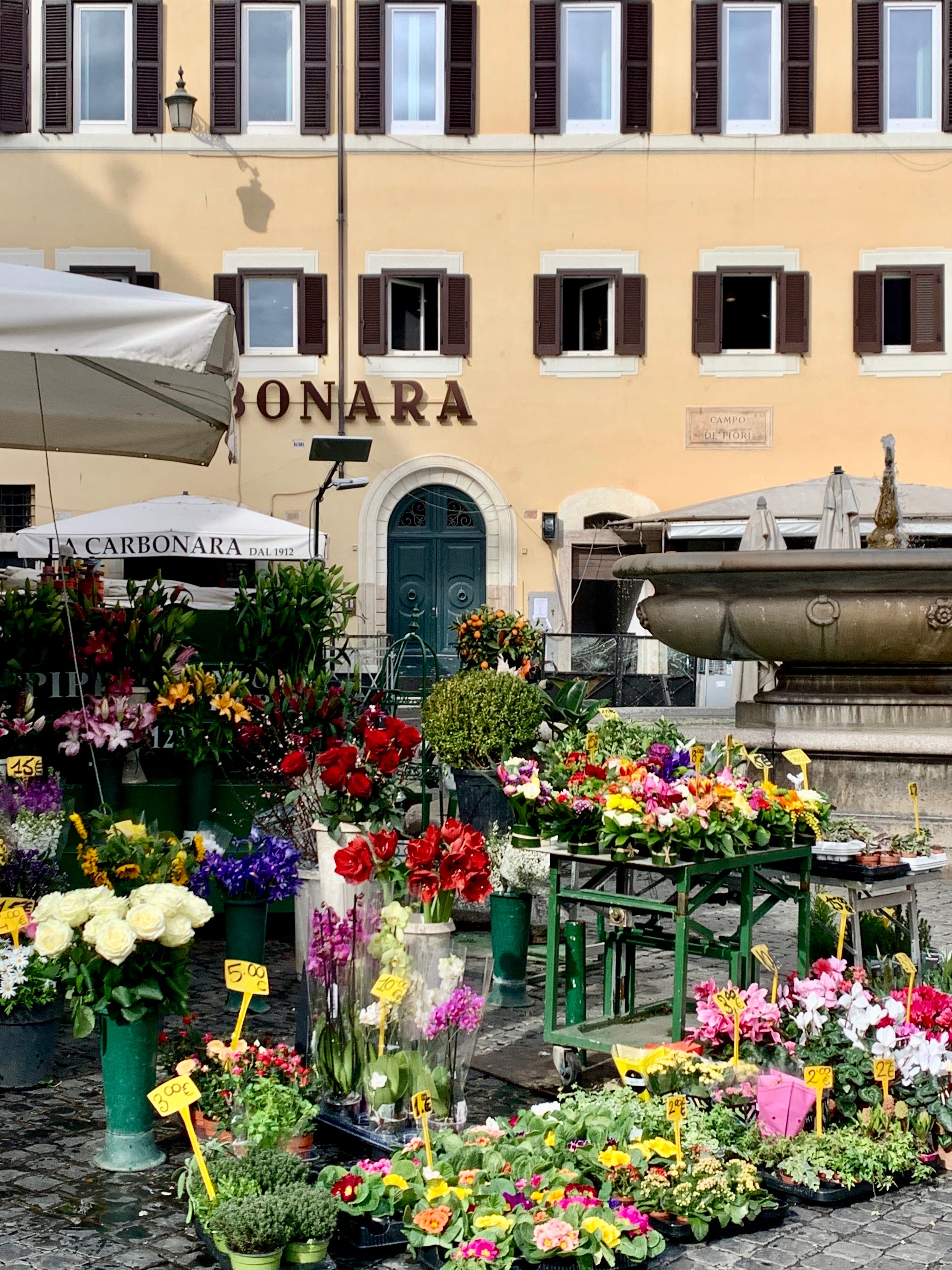 A colorful flower stall with various bouquets and potted plants is set up in an outdoor square in front of a yellow building with brown shutters and a decorative fountain.