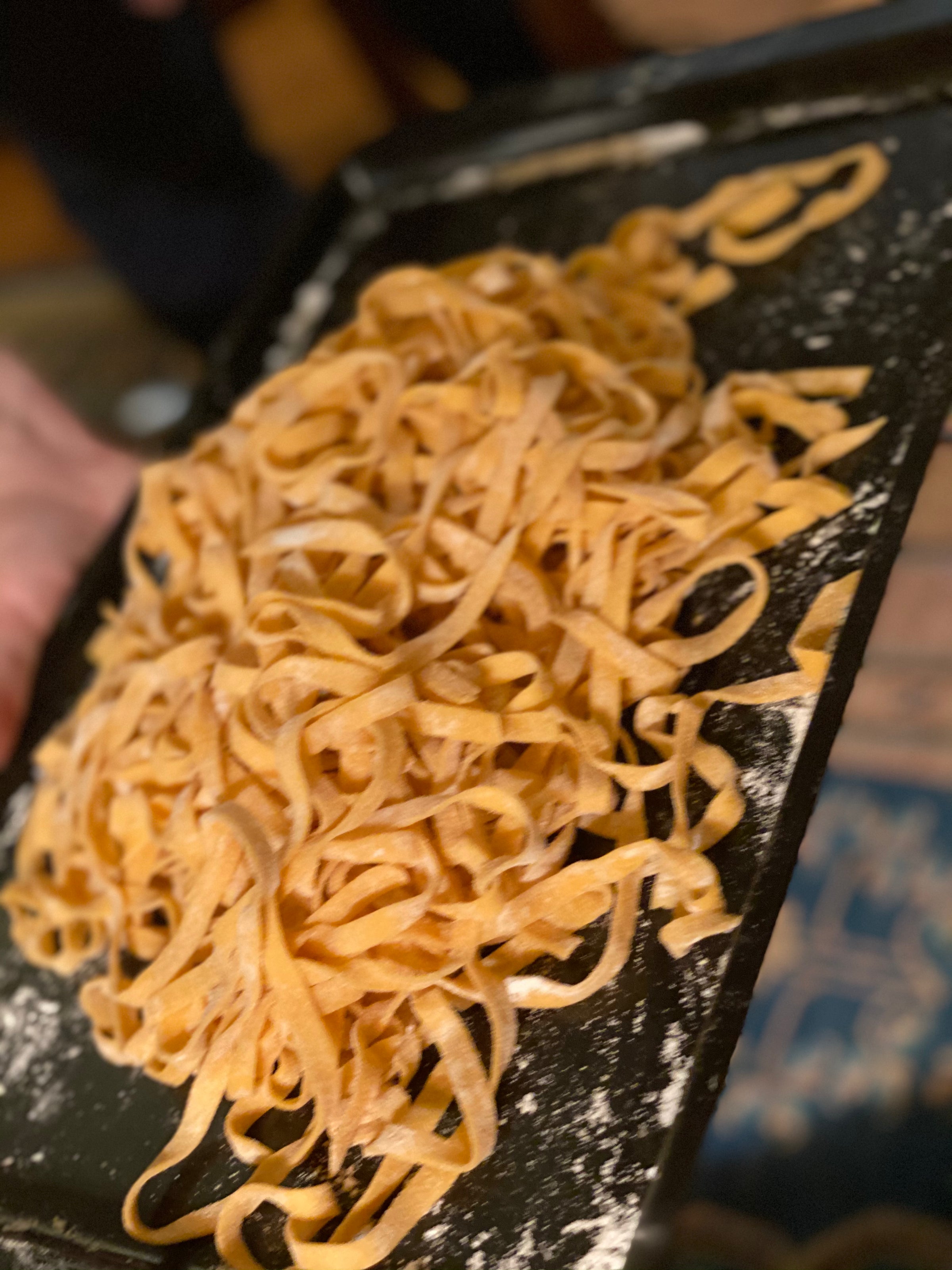 A close-up of a tray filled with fresh, uncooked fettuccine pasta dusted with flour. The noodles are piled loosely on the black tray.