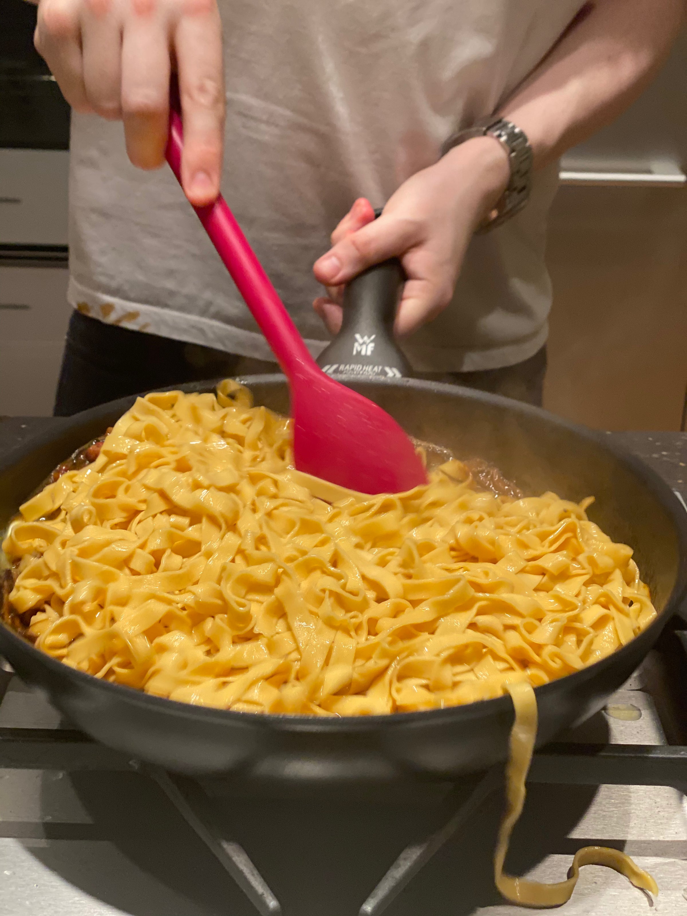 A person in a white shirt stirs a pan of cooked tagliatelle pasta with a pink spatula and a black utensil in a kitchen. Some noodles are hanging over the edge of the pan.