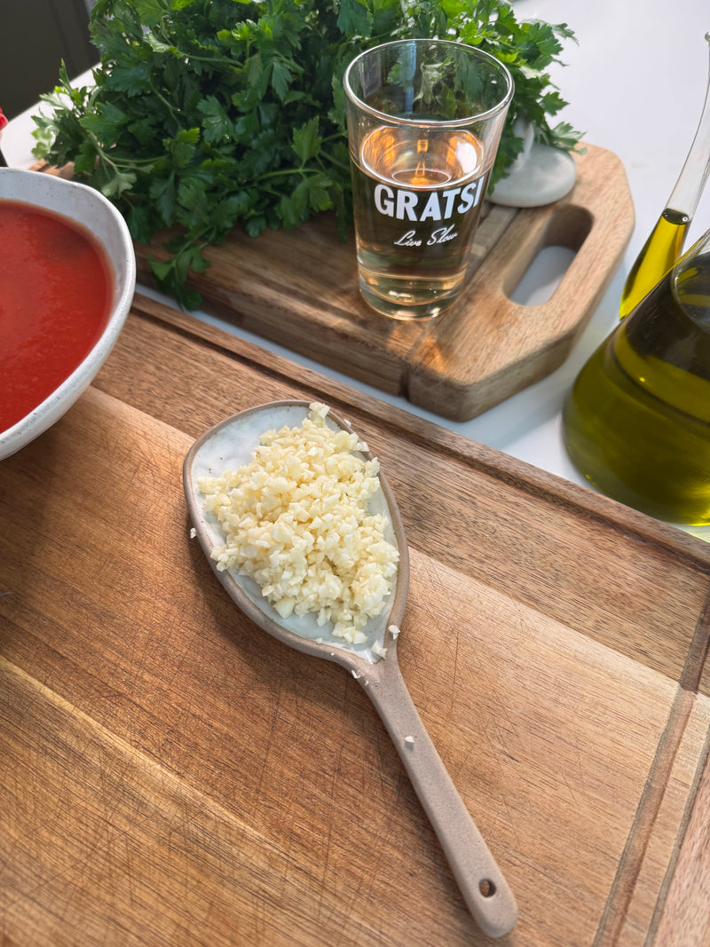 A wooden spoon with chopped garlic rests on a cutting board. Nearby are a bowl of tomato sauce, a glass labeled GRATSI, a bunch of parsley, and bottles of olive oil on a kitchen counter.