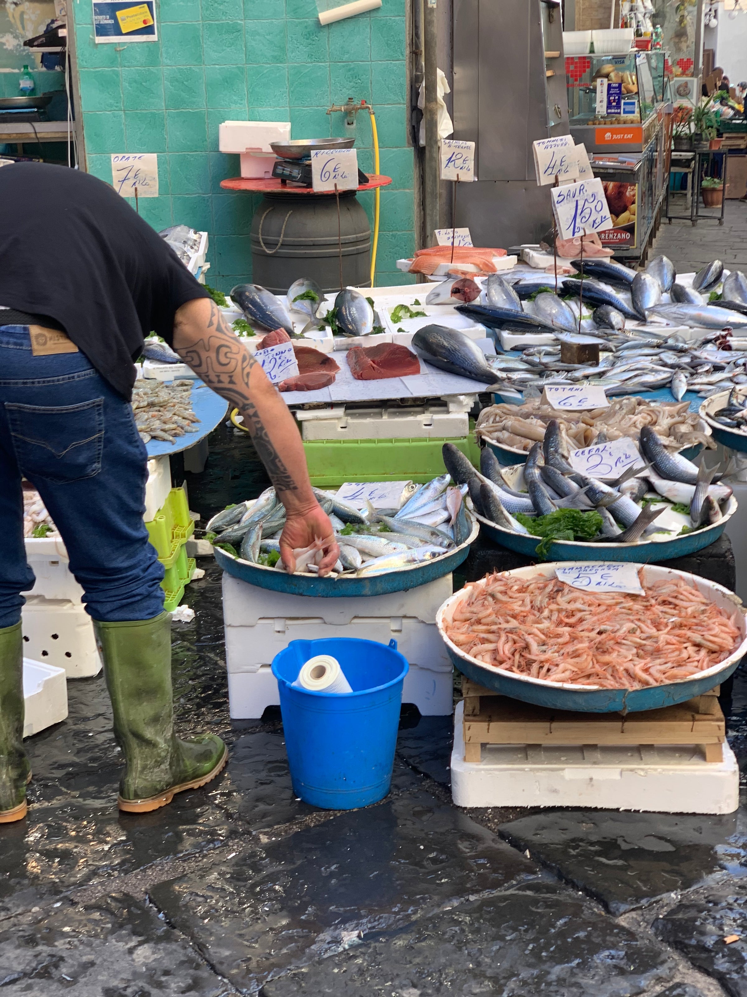 A person wearing green boots selects fish at an outdoor market stall displaying various fresh fish and seafood on ice, with handwritten price signs and a blue bucket on the ground.