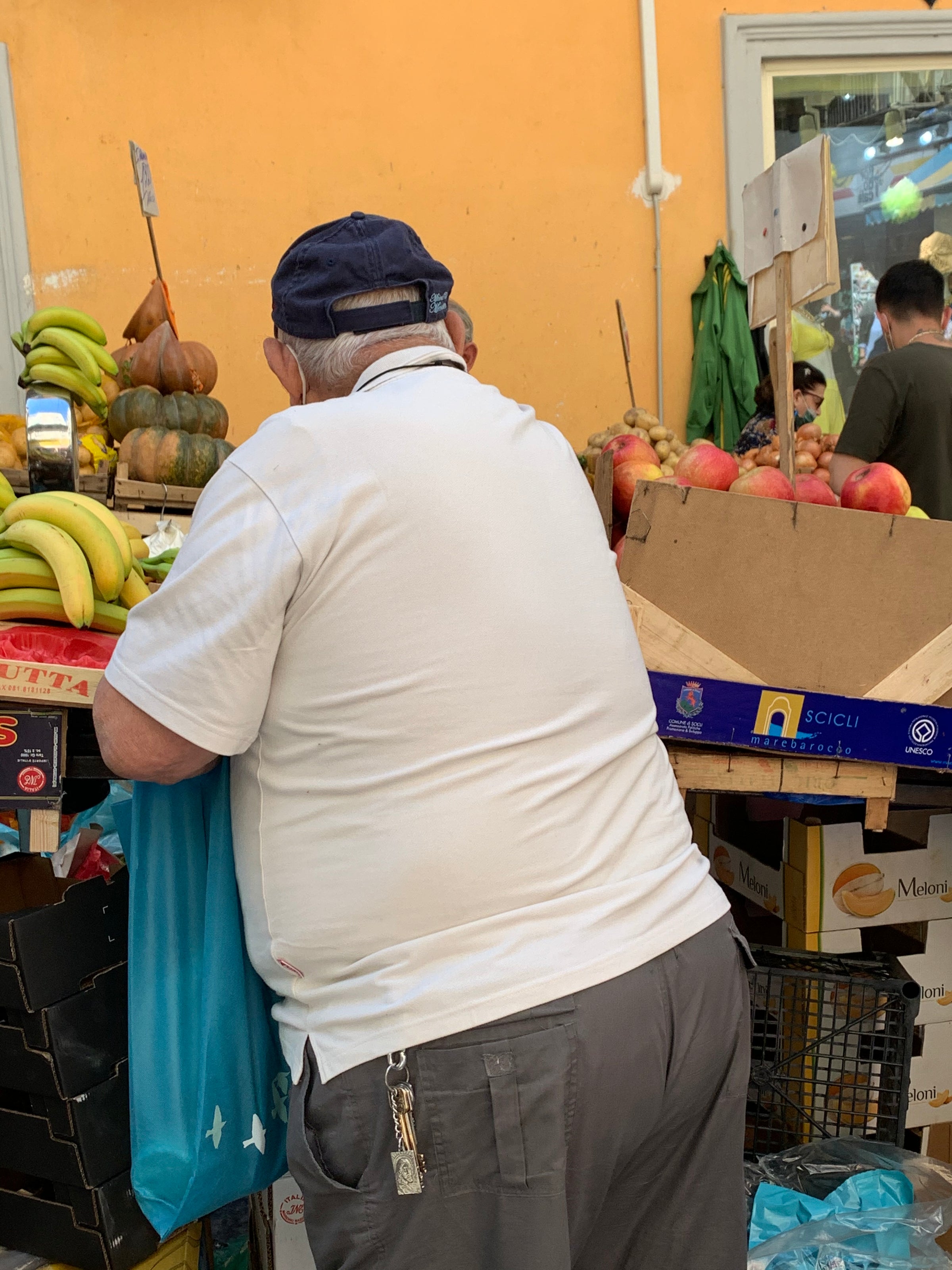 An older man in a white shirt and gray pants shops at an outdoor fruit stand, holding a blue bag. Bananas, apples, and other produce are displayed around him. The scene is lively and colorful.