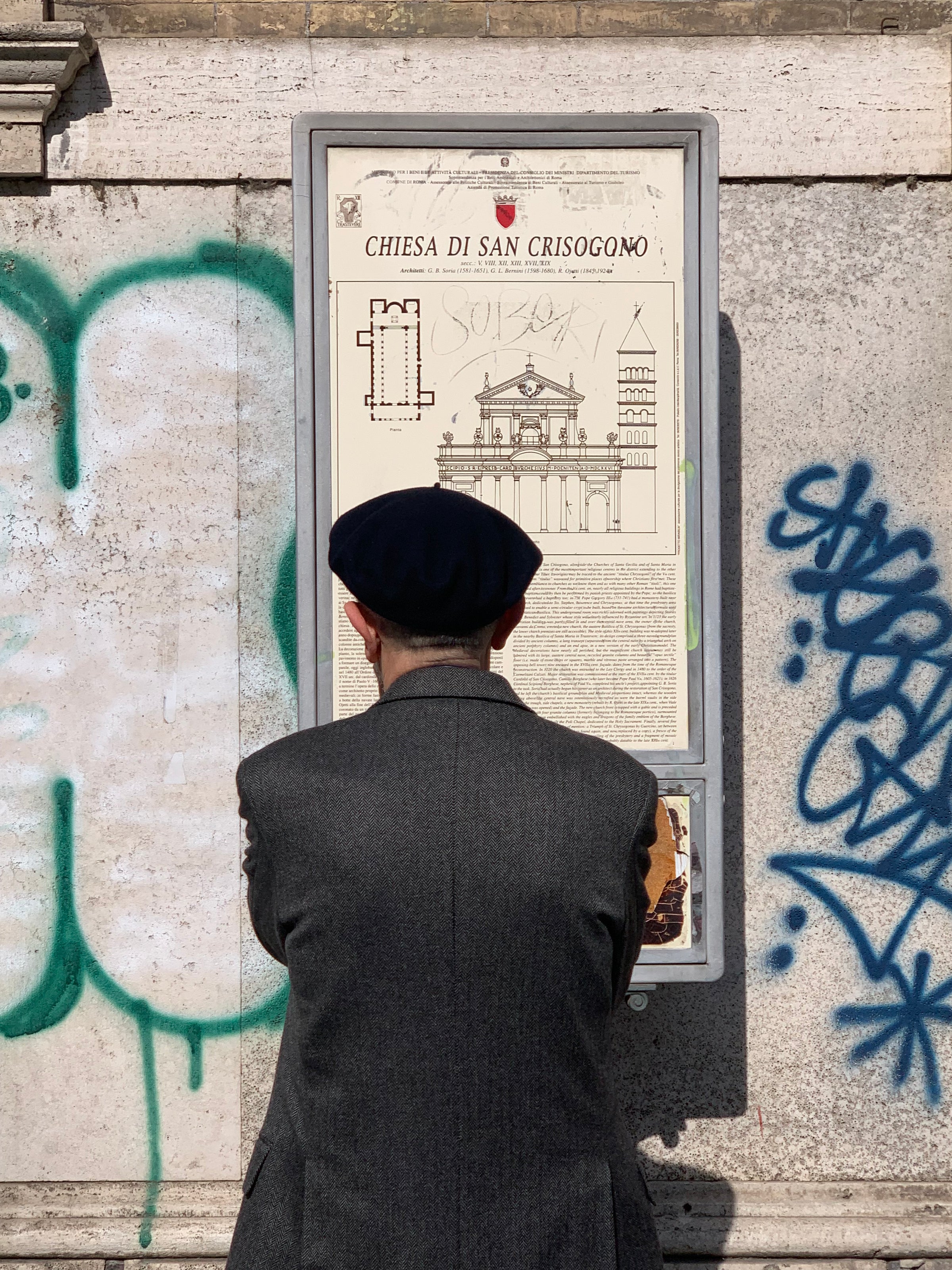 A person wearing a dark beret and gray coat stands facing a sign with an architectural drawing and text titled “Chiesa di San Crisogono” on a stone wall with colorful graffiti.