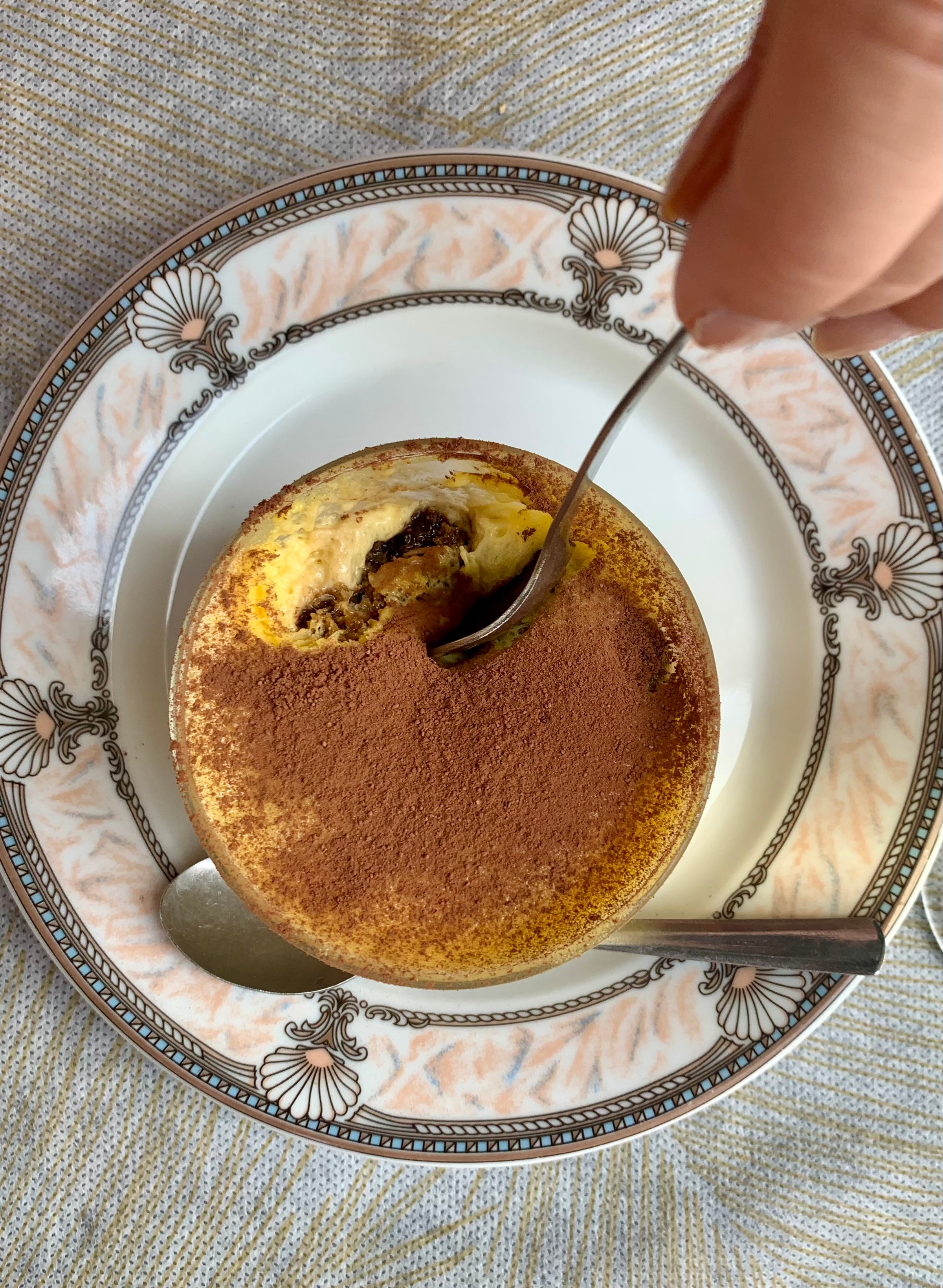 A hand holds a spoon, taking a bite from a round dessert topped with cocoa powder, served on a decorative plate with another spoon resting beside it.