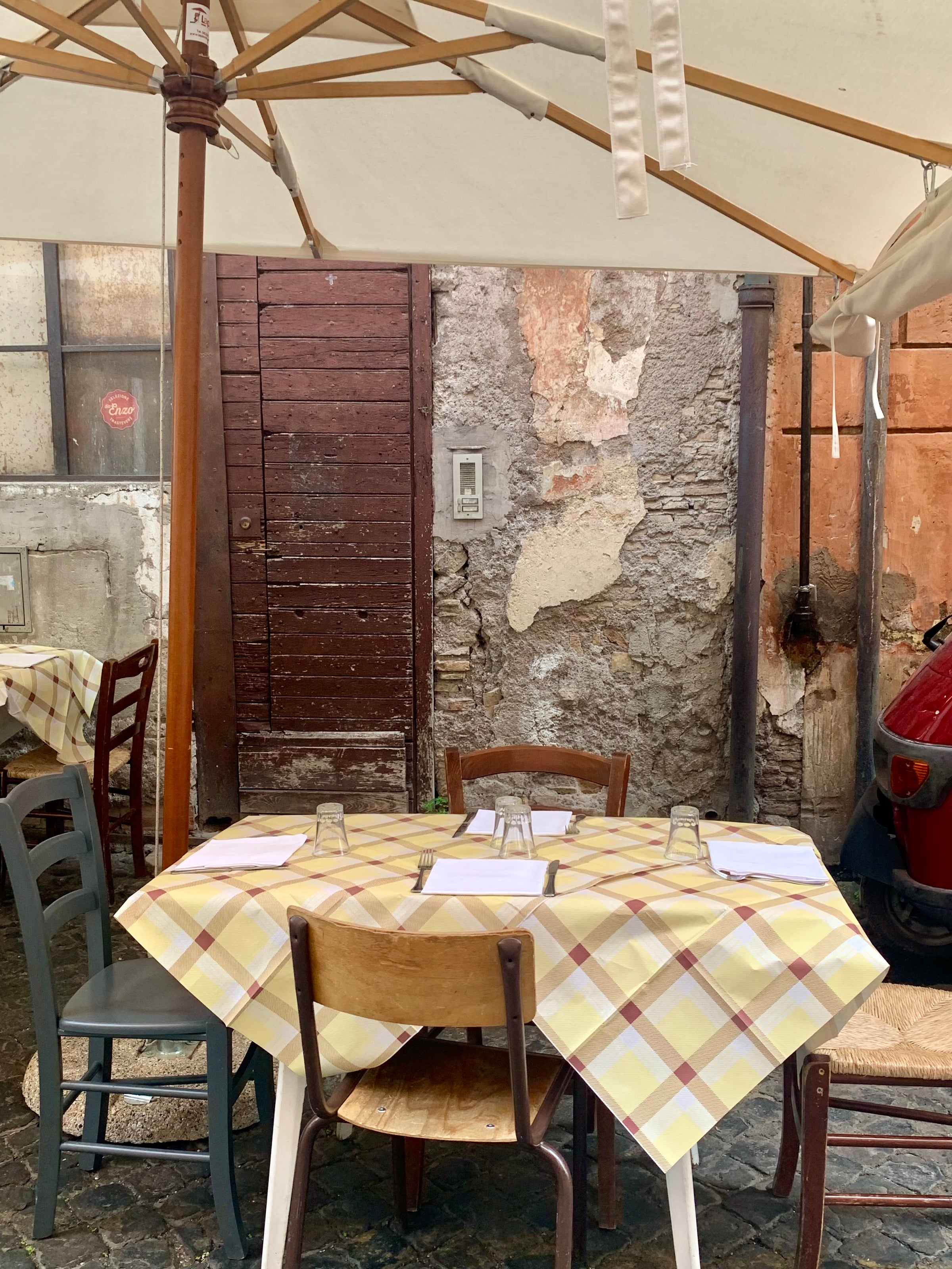 A small outdoor café table with a yellow plaid tablecloth, surrounded by mismatched chairs, set for four with glasses and napkins, under a white umbrella in front of a weathered, rustic wall.