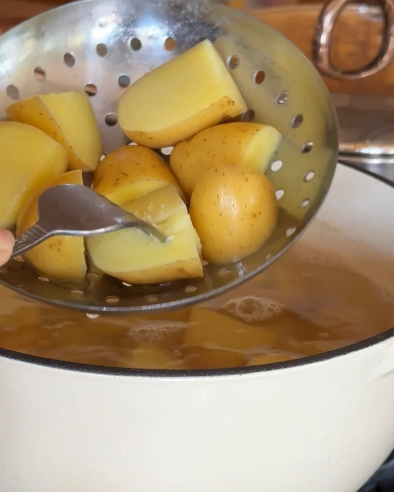 A fork is used to check the tenderness of boiled potato pieces held over a pot of hot water with a slotted spoon.