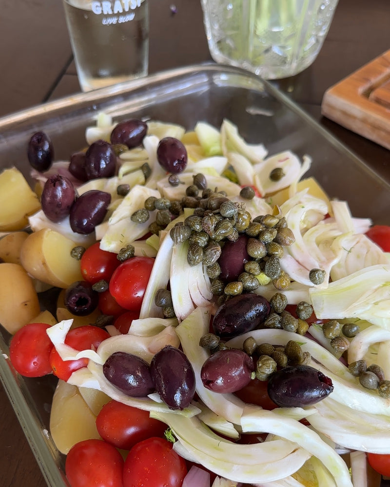 A glass dish filled with a salad of cherry tomatoes, sliced potatoes, fennel, Kalamata olives, and capers. A glass of water and wooden board are visible in the background.