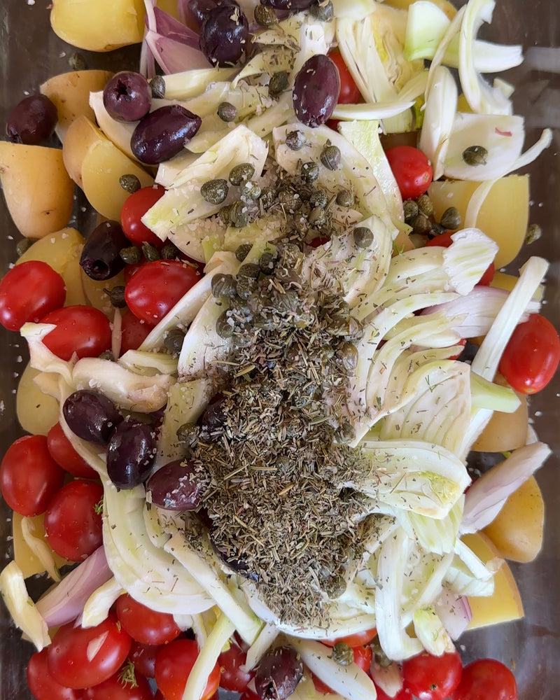 A close-up of a dish with sliced fennel, cherry tomatoes, yellow potato chunks, black olives, capers, and dried herbs, all arranged together and ready to be cooked.