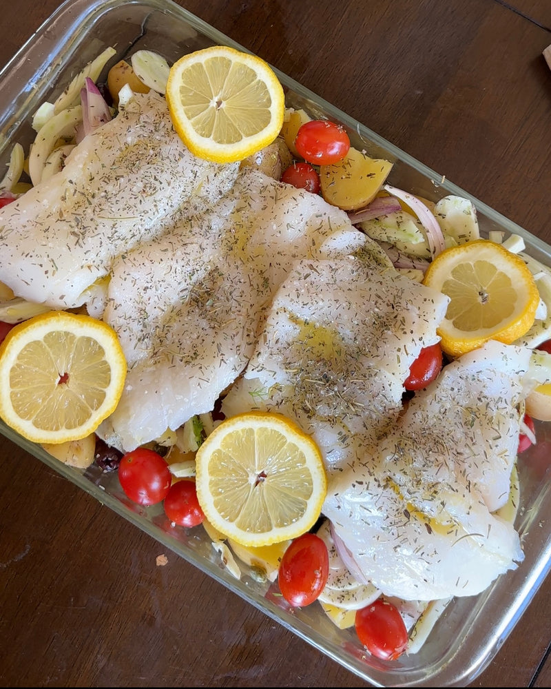 A glass baking dish with raw white fish fillets topped with herbs, surrounded by lemon slices, cherry tomatoes, red onion, and chopped yellow bell pepper, on a wooden table.