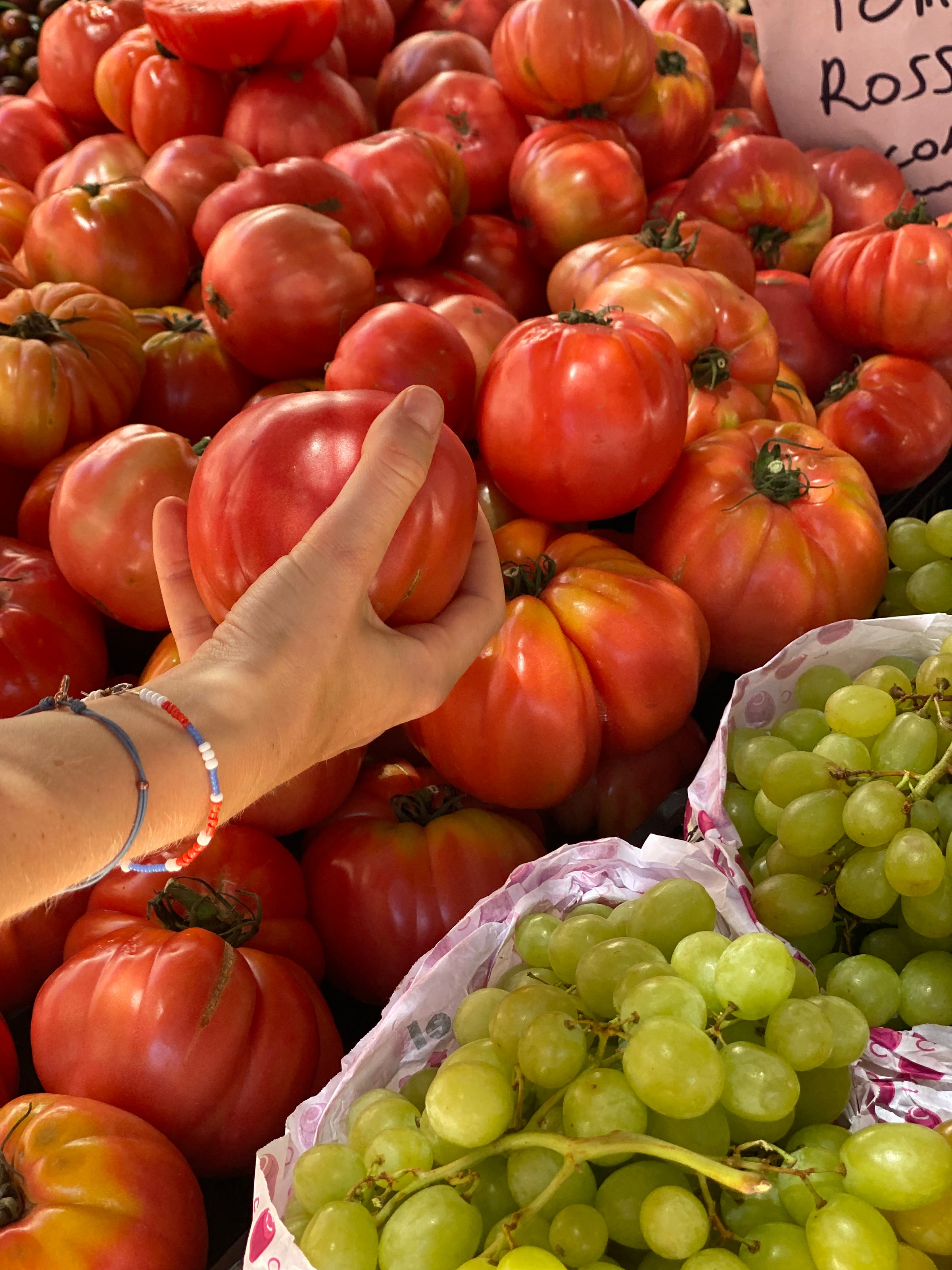 A hand holds a large, ripe tomato over a pile of similar tomatoes at a market, with green grapes in the foreground and a sign partially visible in the background.
