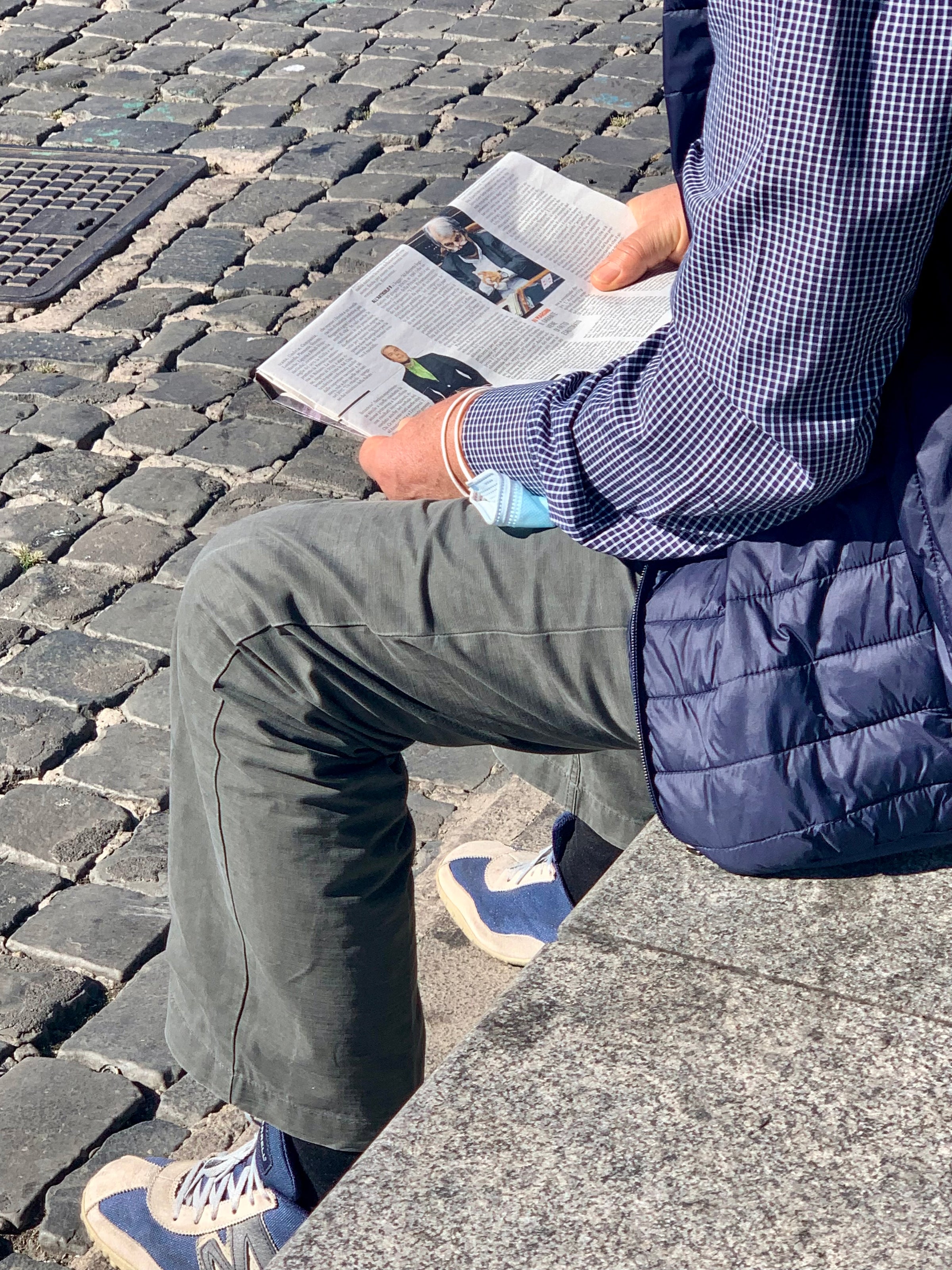 A person wearing a blue checkered shirt, gray pants, and sneakers sits on a stone bench outdoors and reads a magazine. Cobblestone pavement is visible in the background.