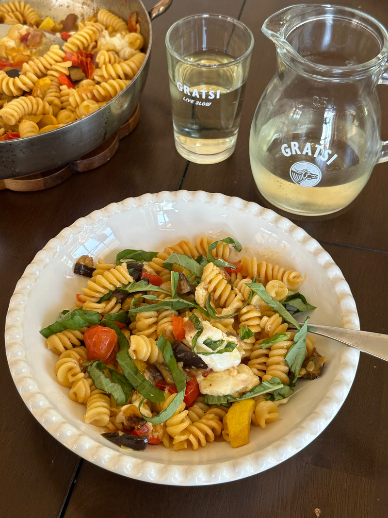 A bowl of rotini pasta with roasted vegetables, fresh basil, and cheese sits on a wooden table, next to a pan of pasta, a glass, and a pitcher of white wine labeled GRATSI.