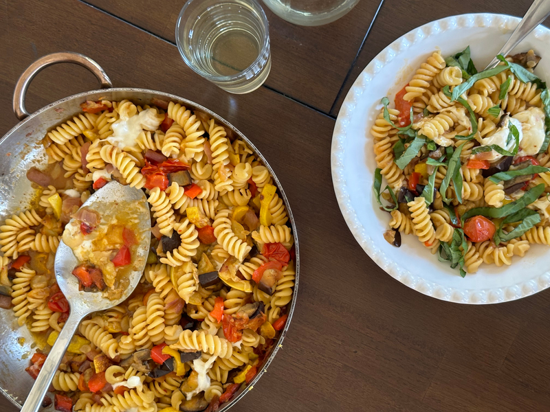 A pan of rotini pasta with tomatoes and vegetables sits next to a white plate of the pasta topped with fresh basil. A glass of water is also visible on the wooden table.