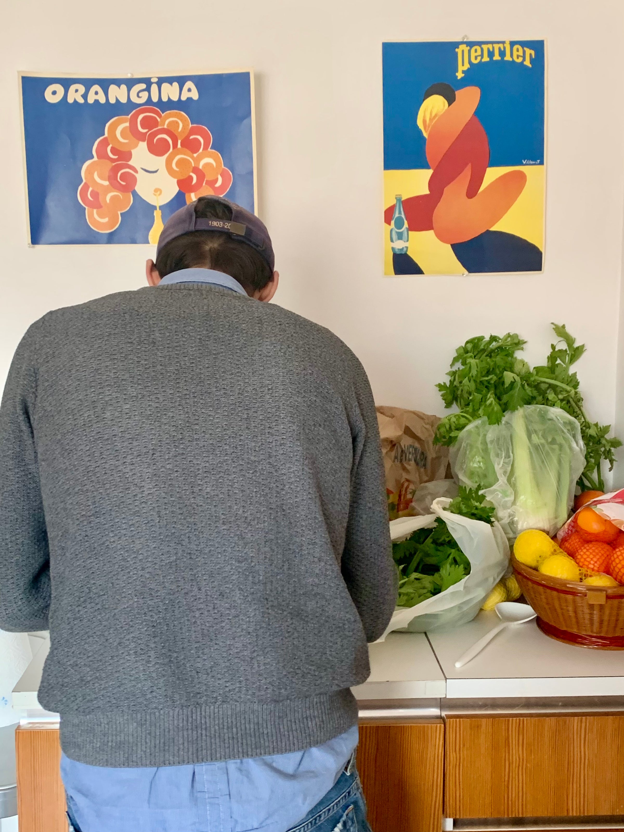A person in a gray sweater and cap stands with their back to the camera at a kitchen counter with fresh produce. Two colorful vintage-style posters for Orangina and Perrier hang on the wall above.