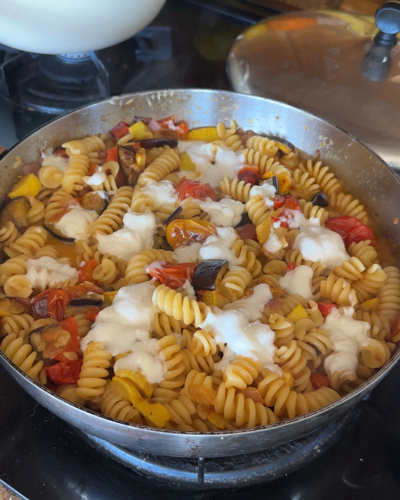 A pan of rotini pasta with roasted cherry tomatoes, eggplant, yellow bell peppers, and melted mozzarella cheese on top sits on a stovetop.