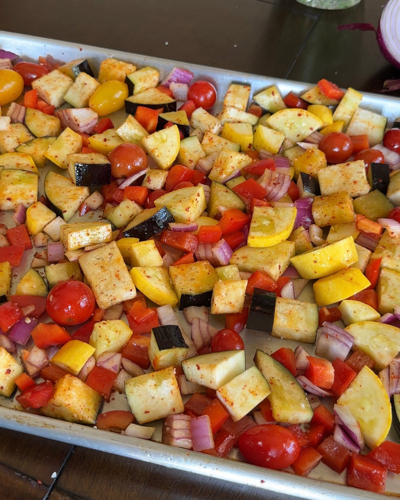 A baking sheet filled with chopped vegetables including yellow squash, eggplant, red bell peppers, red onions, and cherry tomatoes, all seasoned and ready for roasting.