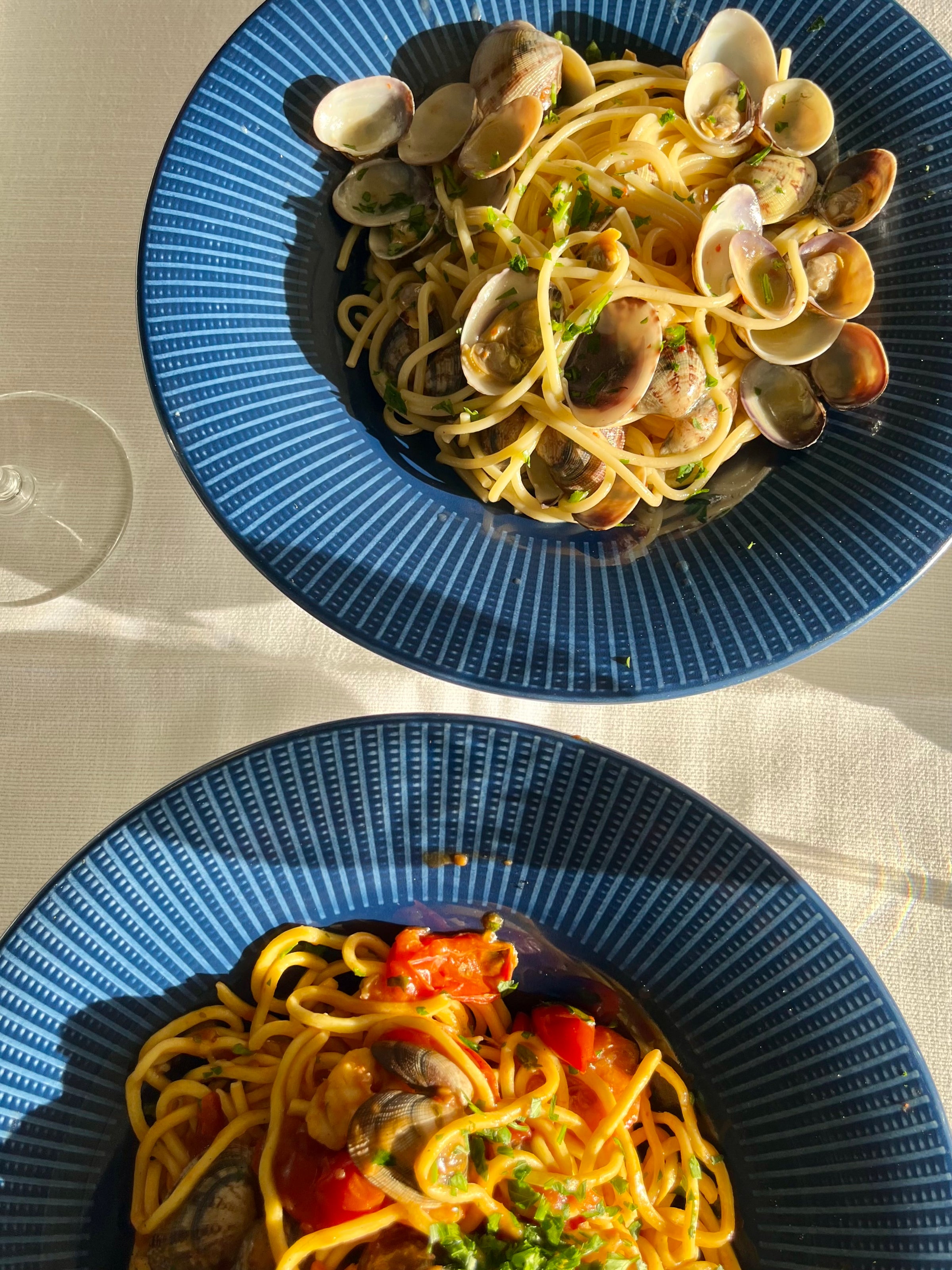 Two blue plates on a white tablecloth, each with a serving of spaghetti; the top plate has spaghetti with clams, while the bottom plate features spaghetti with tomatoes and shellfish. Sunlight casts soft shadows across the table.