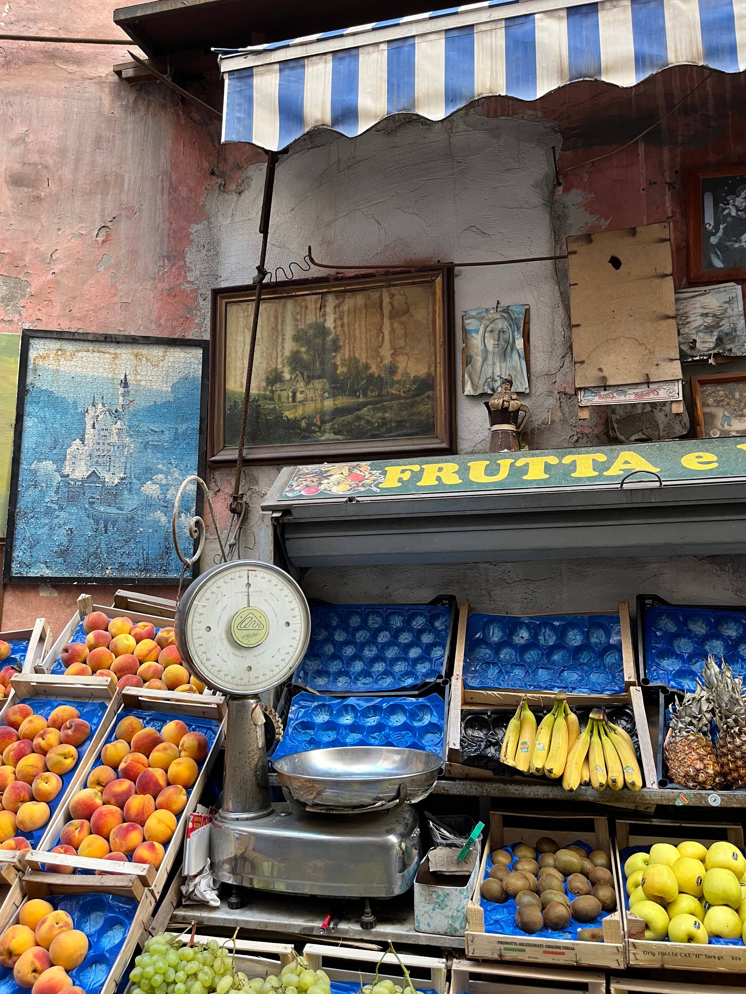 Fruit stall with peaches, bananas, kiwis, and grapes in blue crates; vintage weighing scale in front; striped awning and eclectic wall art above the stand; old, weathered wall in the background.