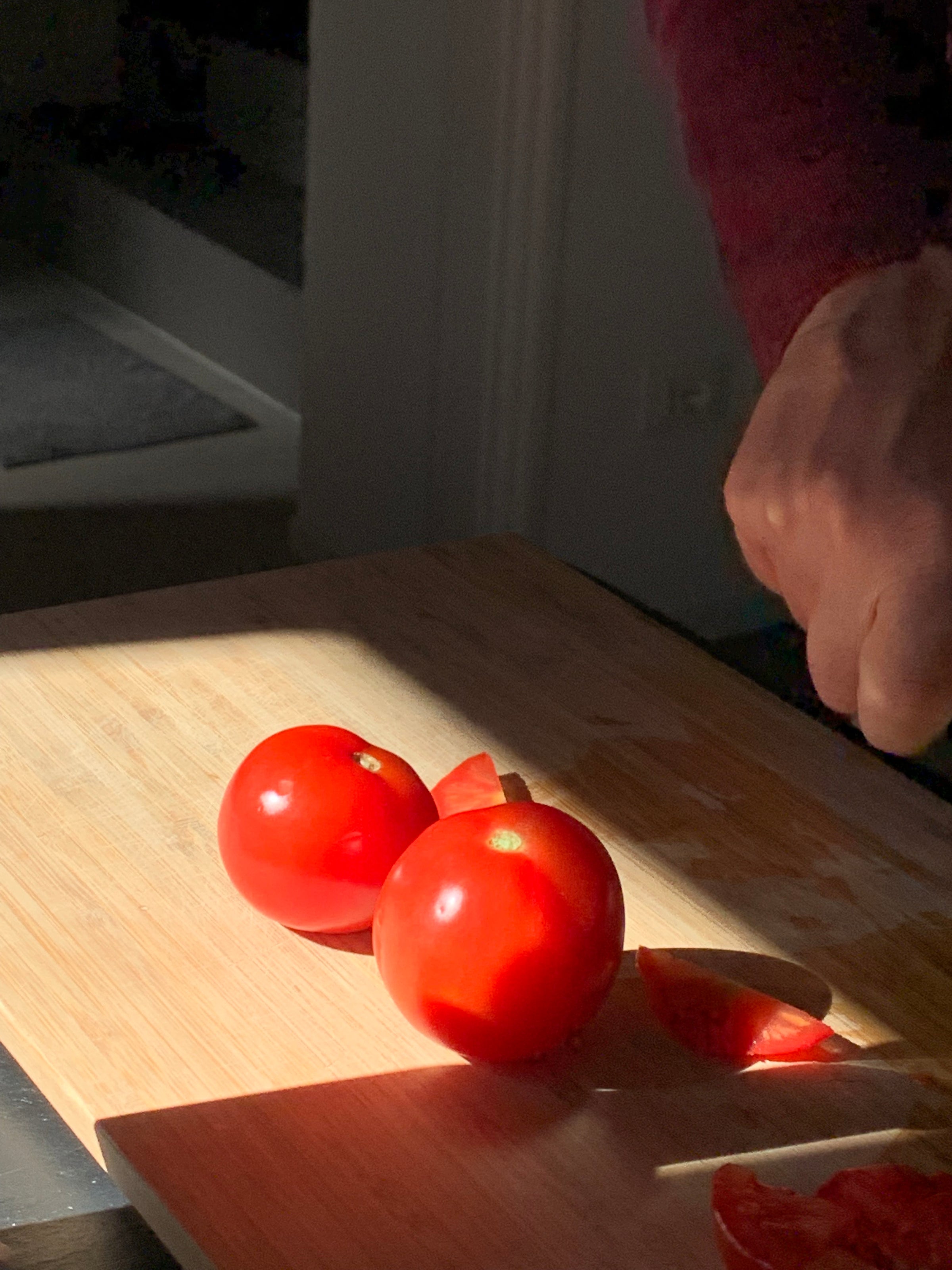 Two whole tomatoes and a few sliced tomato pieces sit on a wooden cutting board, with a person’s hand holding a knife in the process of cutting. Sunlight shines brightly on the tomatoes and board.