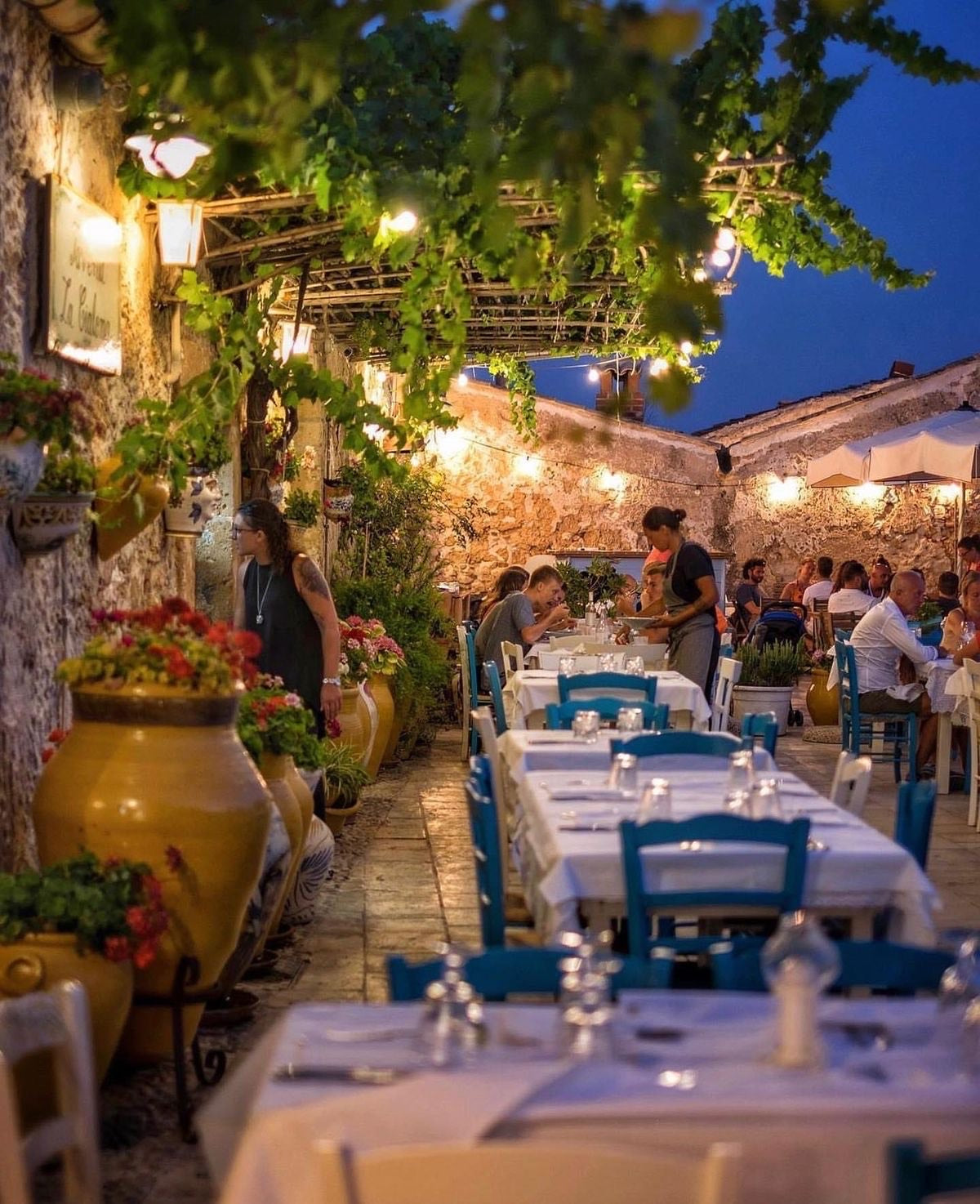 A charming outdoor restaurant with white tablecloths, blue chairs, potted plants, and string lights overhead. Diners enjoy a cozy evening meal under a canopy of vines in a rustic, stone-walled courtyard.