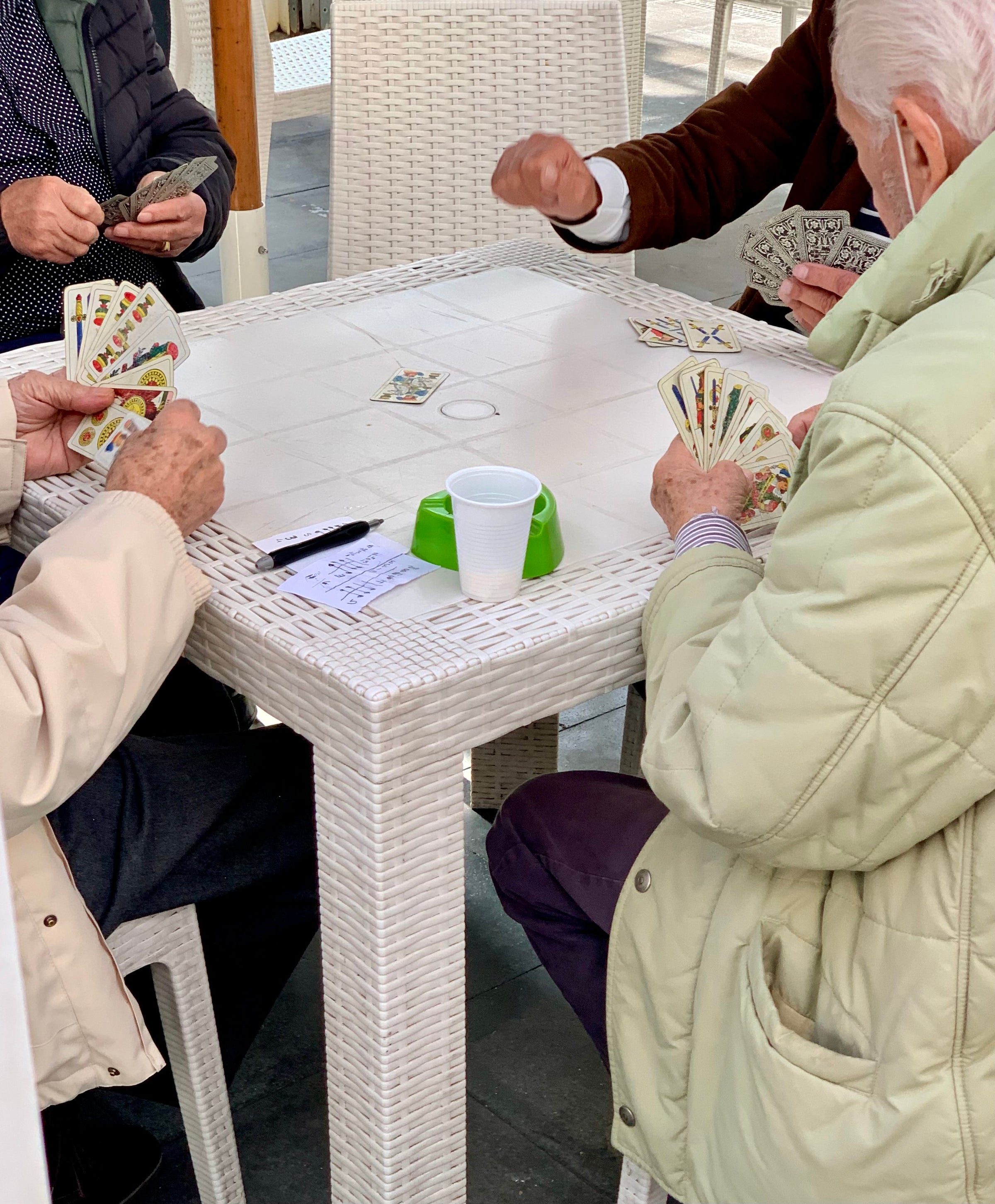 Four elderly people sit around a white outdoor table playing cards. Their faces are not visible, and a white cup and a pen with a small notepad are on the table. The scene suggests a relaxed social gathering.