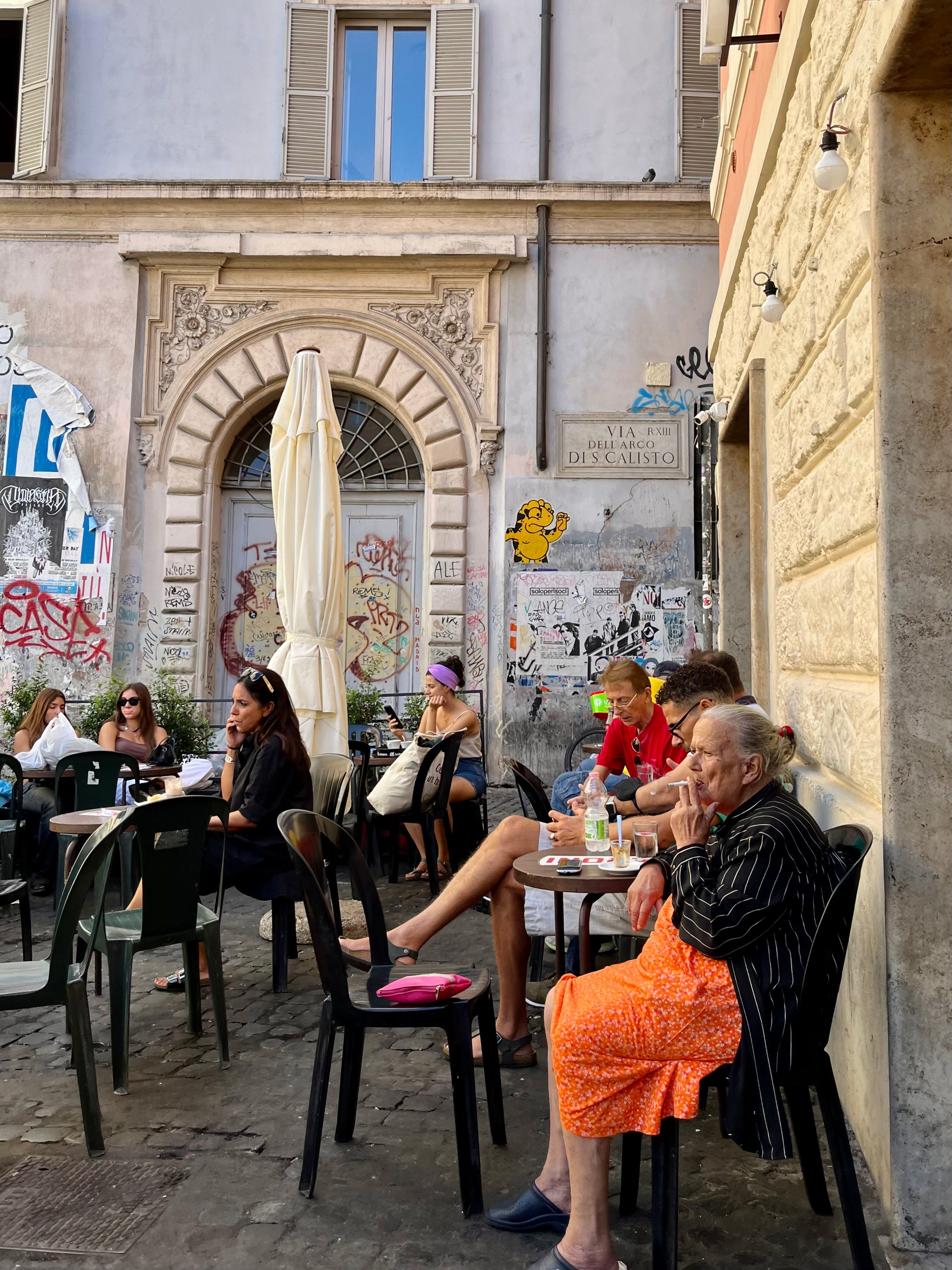 People sit at outdoor café tables beside a graffiti-covered wall in a European city. An elderly woman in an orange dress is in the foreground, and others relax and chat in the sun. The setting is lively and casual.