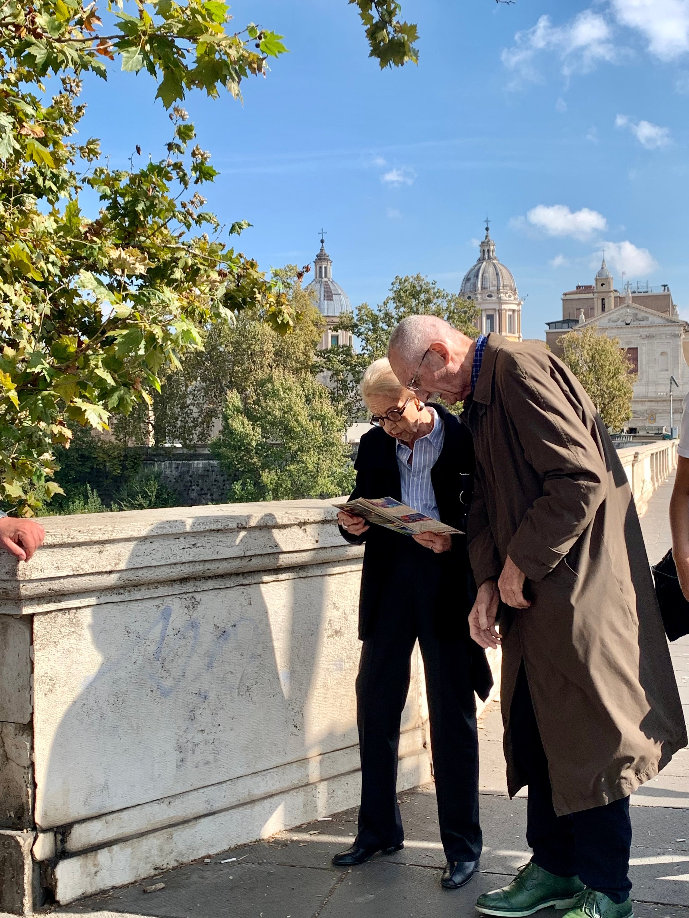 Two elderly men stand on a sunny stone bridge, one holding and reading a map. Trees and historic buildings with domes are visible in the background under a blue sky.