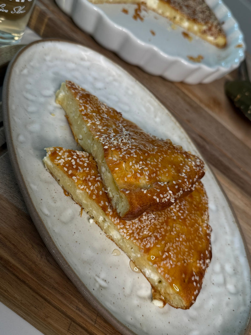 Two triangular slices of golden, sesame-topped baked cheese pie are served on a white ceramic plate, with a round baking dish containing more pie visible in the background.