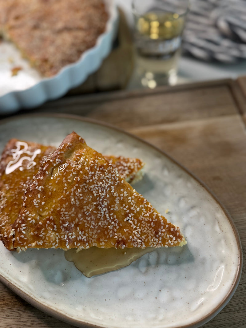Two triangular slices of sesame-topped bread drizzled with honey sit on a ceramic plate, with a pie dish and a glass blurred in the background on a wooden surface.