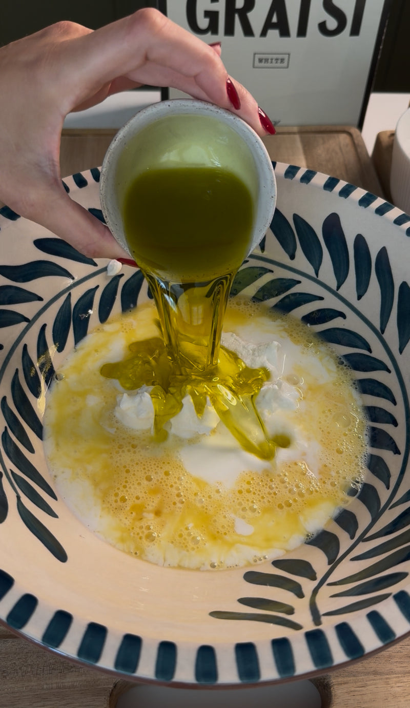 A hand pours greenish olive oil from a small cup into a bowl containing whisked eggs, milk, and possibly flour, preparing ingredients for baking or cooking.