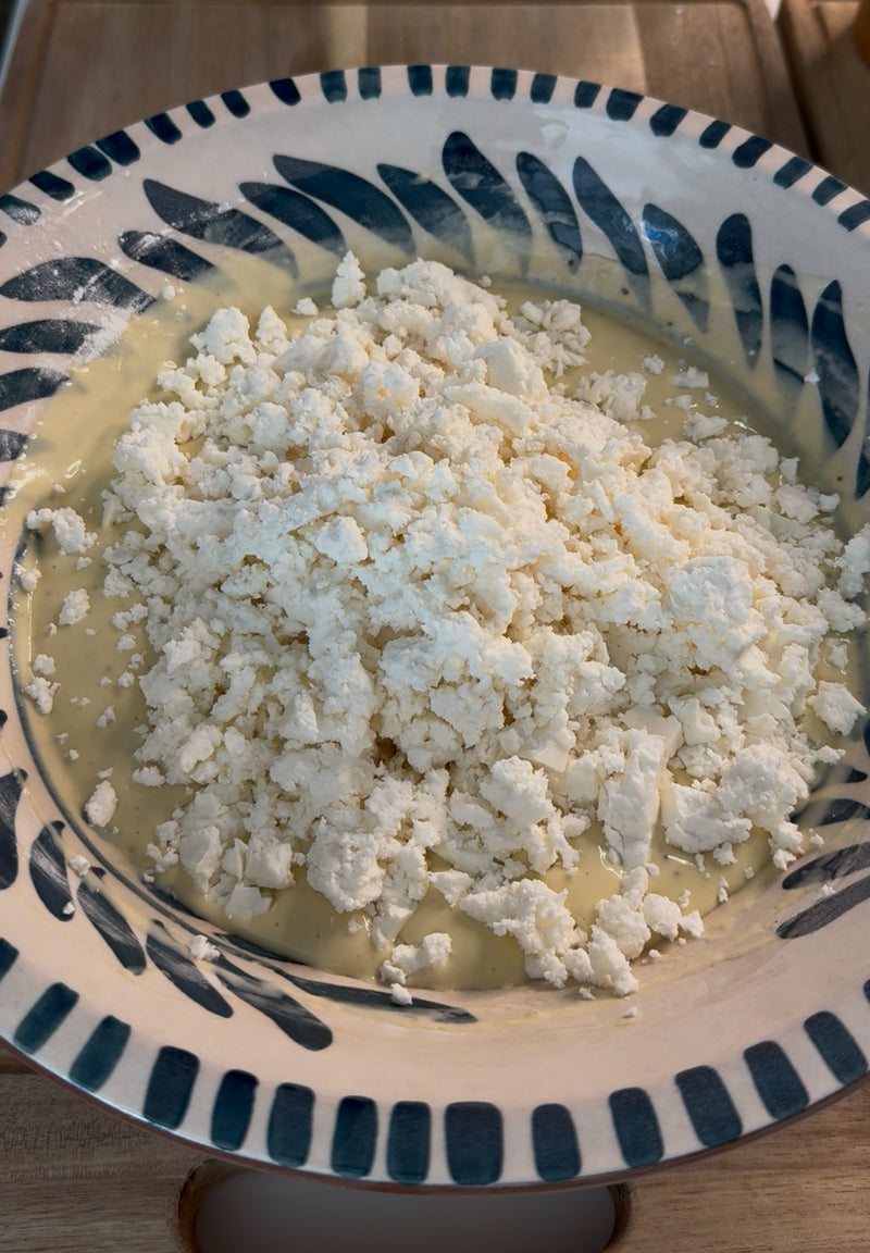 A patterned bowl filled with a creamy yellow mixture, topped with a generous amount of crumbled white cheese, sits on a wooden surface in natural light.