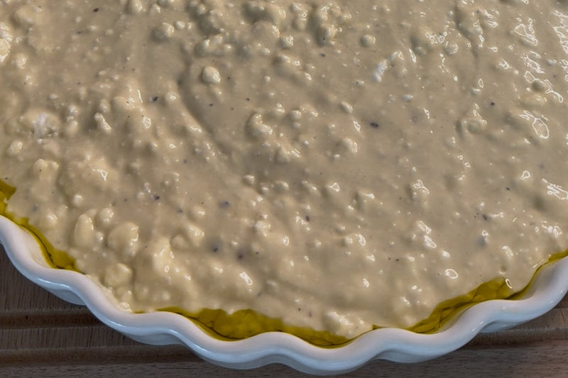 A close-up of an unbaked pie or tart in a white ceramic dish, filled with a lumpy, creamy mixture over a crust, ready for baking. The edge of the crust is visible around the dish.
