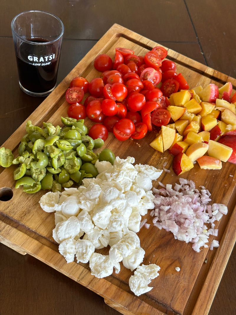 A wooden cutting board holds chopped green olives, cherry tomatoes, peaches, shallots, and mozzarella balls. Next to the board is a glass of red wine with GRATS! printed on it.