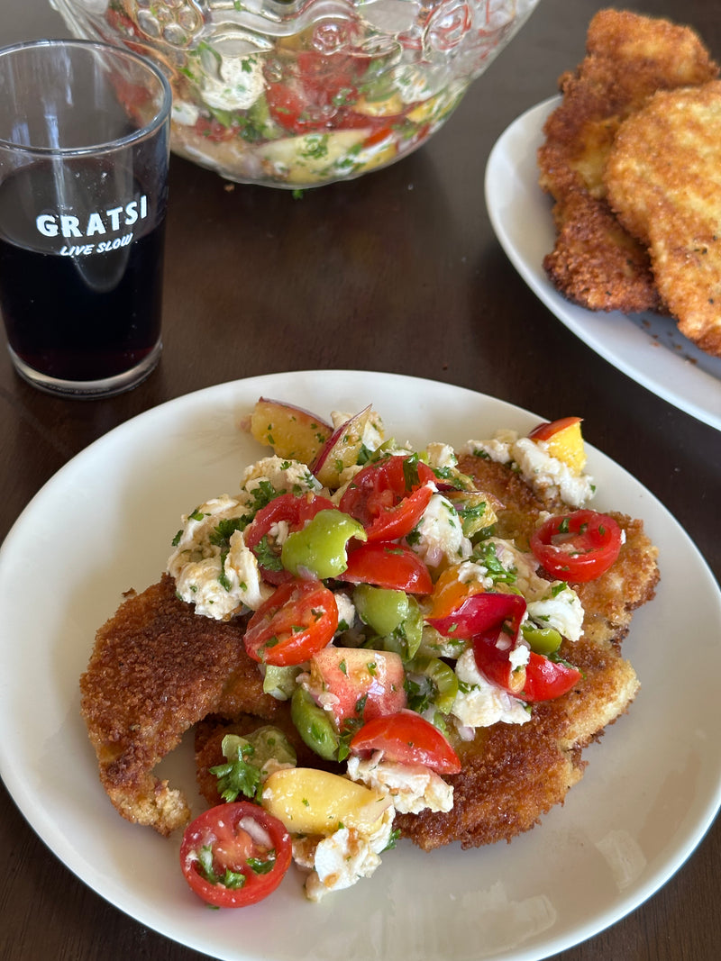 A plate of breaded, fried cutlets topped with a colorful tomato, peach, olive, and cheese salad. In the background are a bowl of salad, a stack of cutlets, and a glass with the word GRATSI.