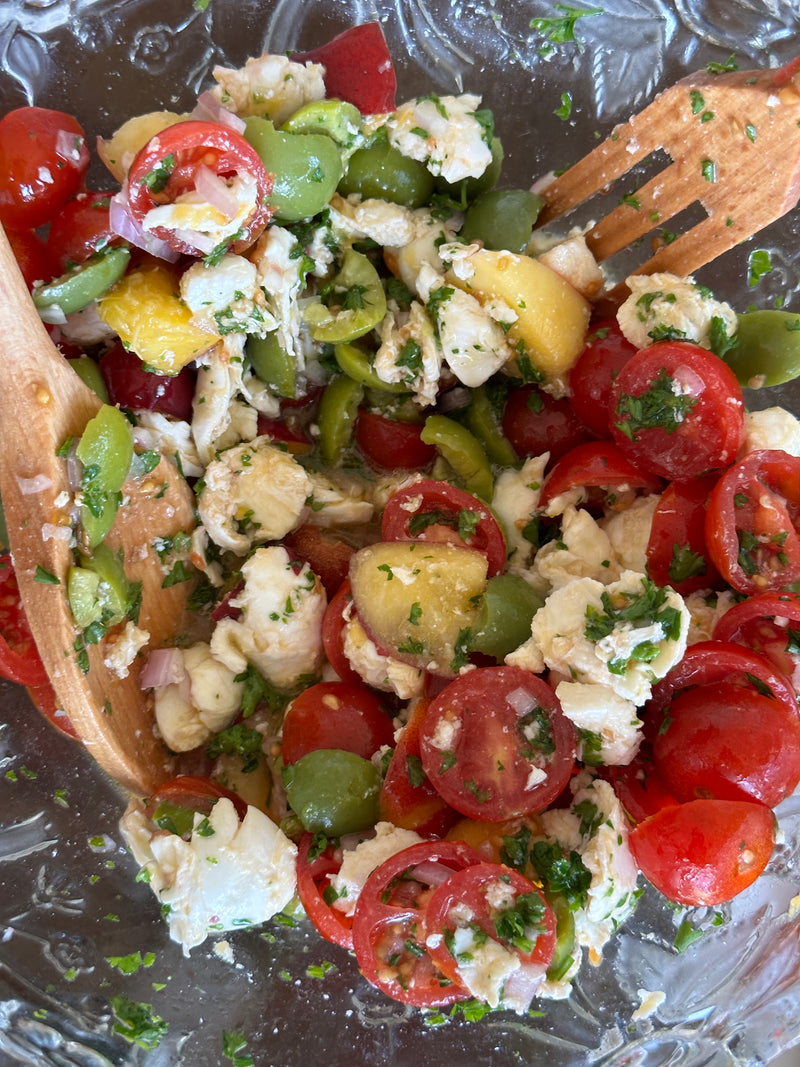 A close-up of a fresh salad with cherry tomatoes, green olives, mozzarella cheese, herbs, and wooden salad tongs in a glass bowl.