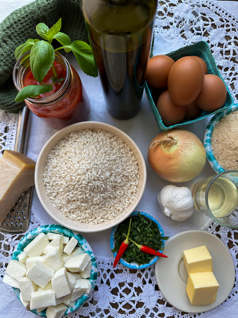 A top view of risotto ingredients on a lace tablecloth: a bowl of rice, eggs, onion, garlic, butter, Parmesan, feta, fresh basil, two red chilies, tomato sauce, breadcrumbs, white wine, and a bottle of olive oil.