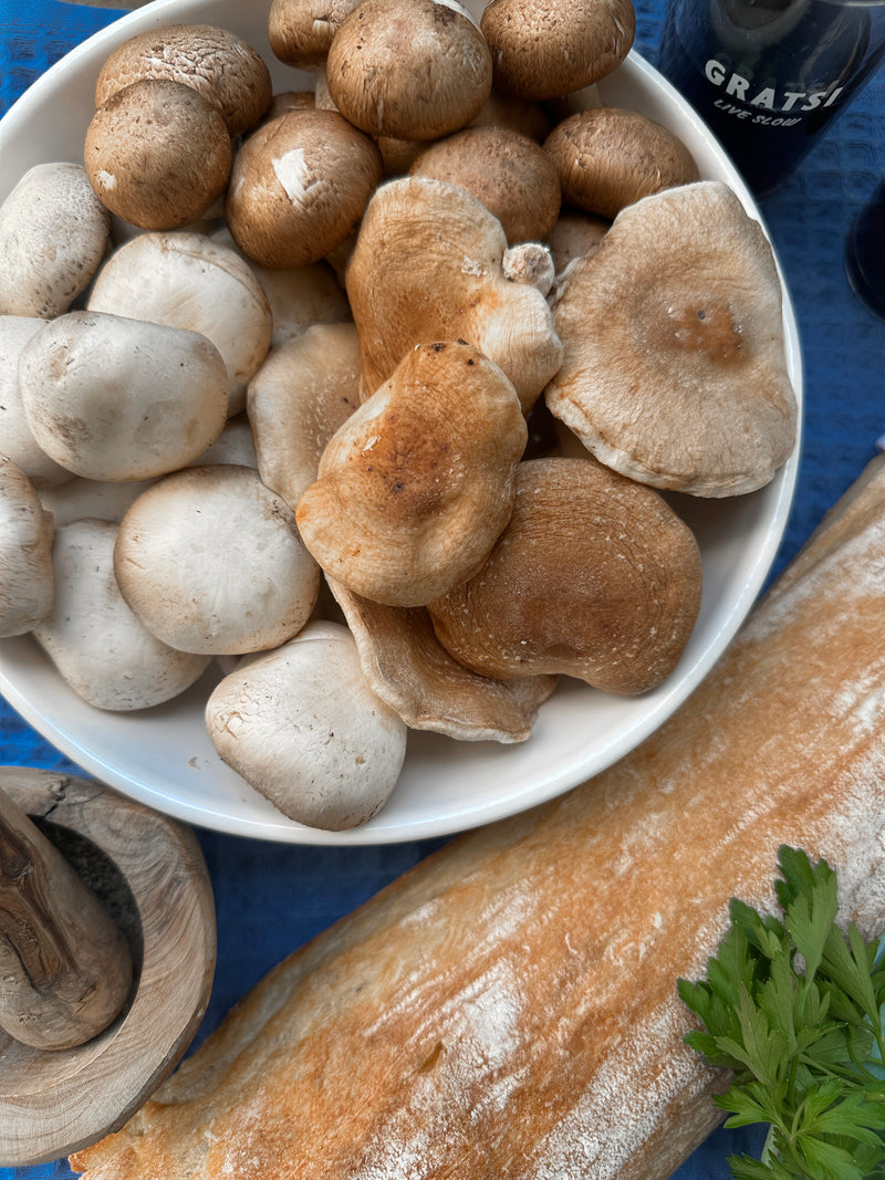 A white bowl filled with various mushrooms sits on a blue surface next to a fresh baguette, a sprig of parsley, a wooden pepper grinder, and a bottle of wine partially visible.