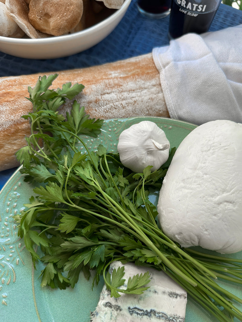A plate with fresh parsley, a head of garlic, a block of mozzarella cheese, and a piece of blue cheese. In the background, there’s a baguette, a bowl of bread rolls, and a dark bottle on a blue cloth.