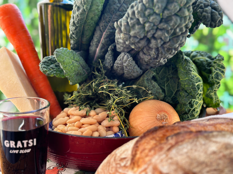 A close-up of vegetables and food items, including kale, a carrot, an onion, white beans, a wedge of cheese, a loaf of bread, olive oil, and a glass of red wine labeled GRATSI LIVE SLOW, arranged on a table.