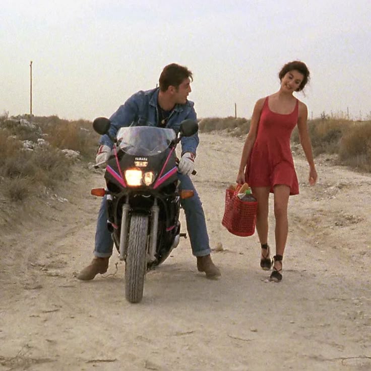 A man on a black Yamaha motorcycle looks at a woman in a red dress walking beside him on a dirt road. She is smiling and holding a red basket. The landscape around them is dry and sparse.